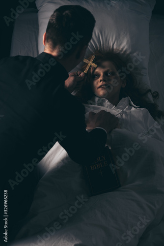 exorcist with bible and cross standing over obsessed girl in bed