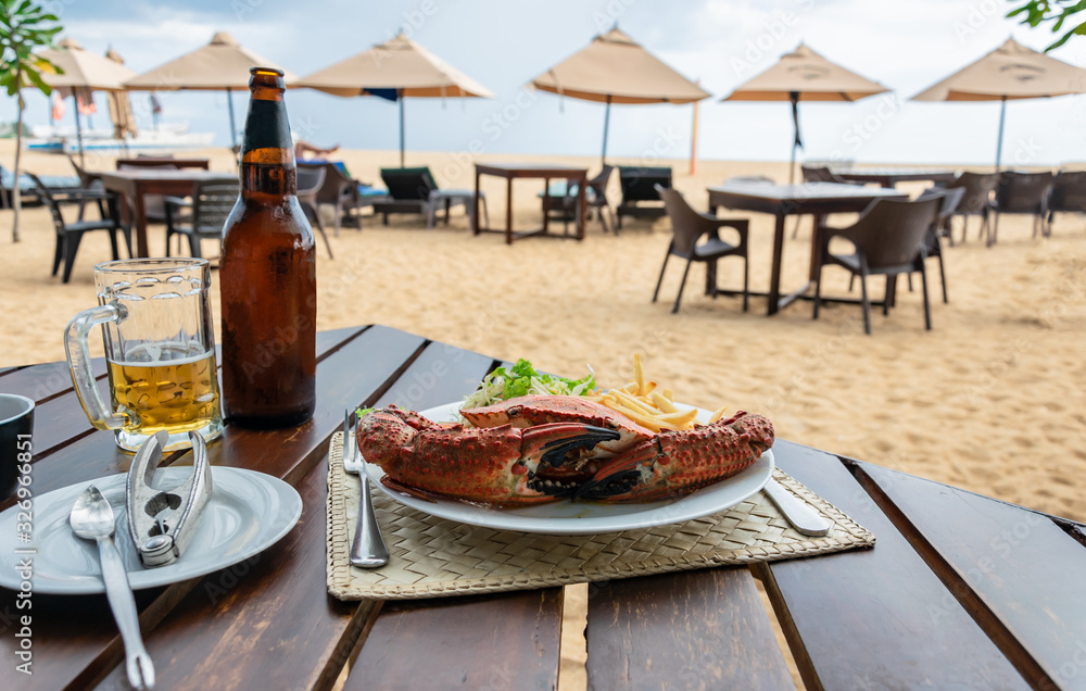 Lunch in a beach restaurant with boiled crab shell, french fries, salad on a white plate and cold light beer, sandy beach on the background
