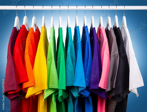row of many fresh new fabric cotton t-shirts in colorful rainbow colors hangng on clothes rail in wrdrobe. Various colored shirts blue white background. diy printing fashion concept.