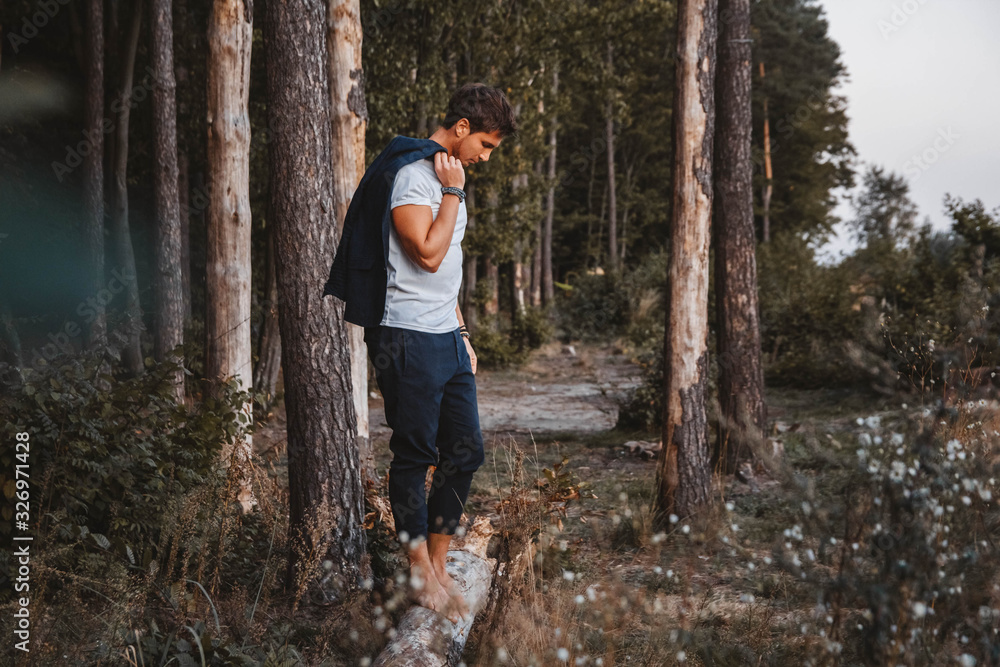 Handsome man posing outdoors in forest stands barefoot on a log, wearing checked jacket. Place for text or advertising Handsome man posing outdoors in forest stands barefoot on a log, wearing checked jacket. Place for text or advertising