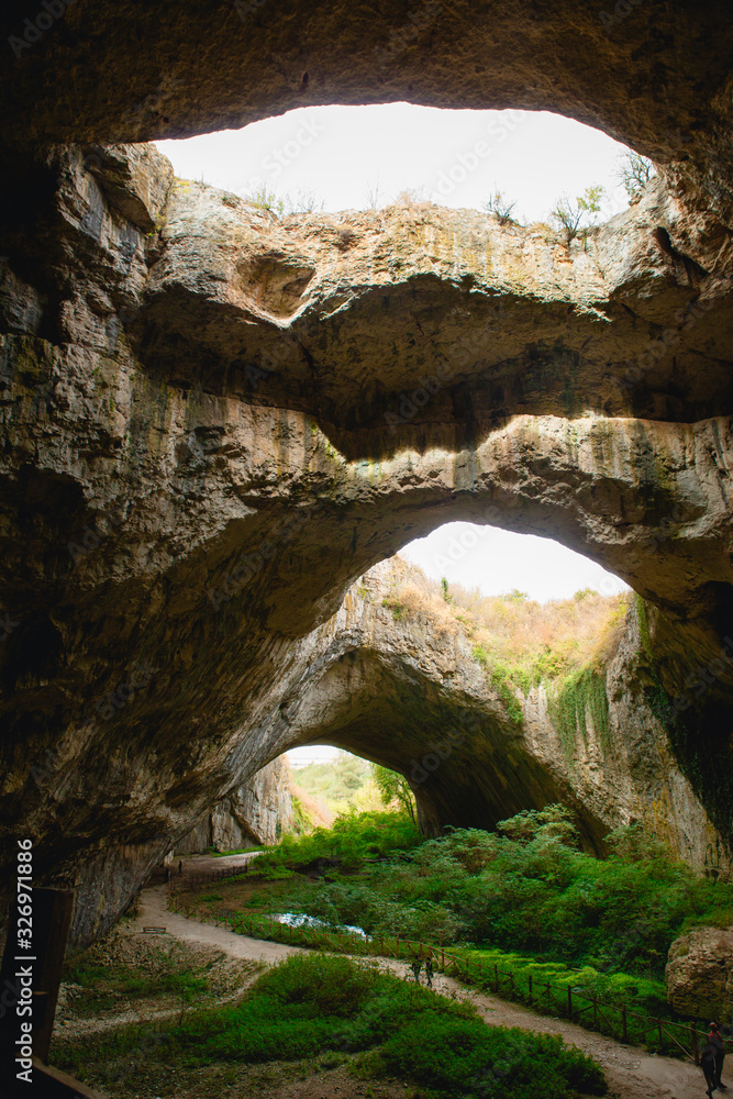 High arches of a huge stone cave with round holes at the top, a tourist road with a fence inside the cave. Bulgaria's natural attraction-Devetashka cave