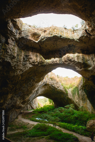 Fototapeta Naklejka Na Ścianę i Meble -  High arches of a huge stone cave with round holes at the top, a tourist road with a fence inside the cave. Bulgaria's natural attraction-Devetashka cave