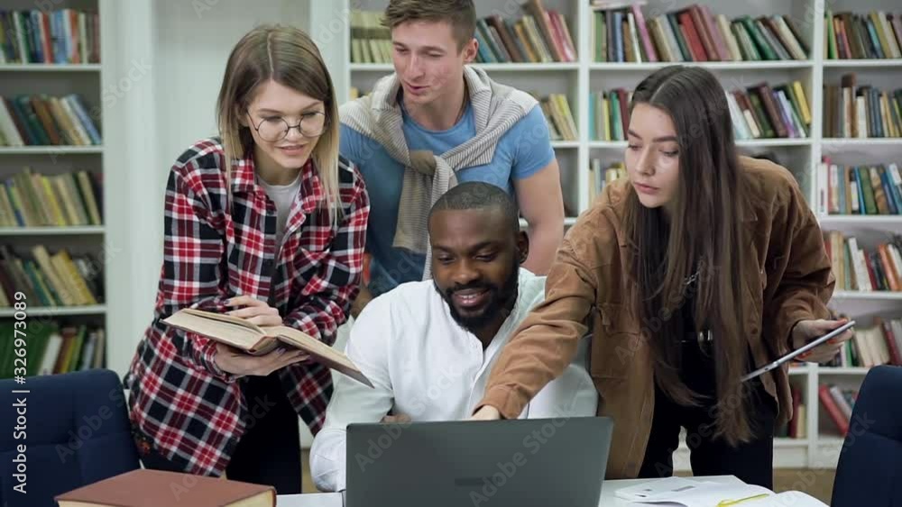 Front view of good-looking happy modern smart multiracial youth which studying in university library using information on laptop Front view of good-looking happy modern smart multiracial youth which studying in university library using information on laptop