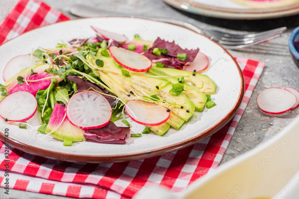 selective focus of fresh radish salad with greens and avocado served on napkin with cutlery on grey concrete surface selective focus of fresh radish salad with greens and avocado served on napkin with cutlery on grey concrete surface