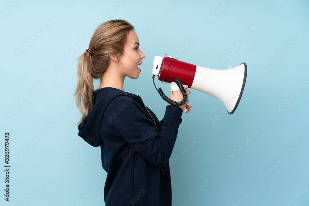 Teenager Russian girl holding mat isolated on blue background shouting through a megaphone Teenager Russian girl holding mat isolated on blue background shouting through a megaphone