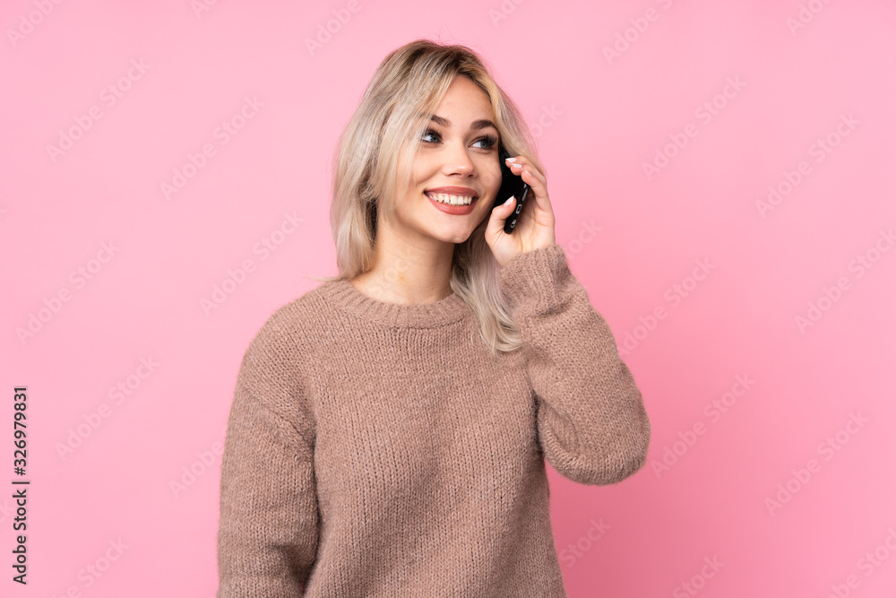 Teenager blonde girl wearing a sweater over isolated pink background keeping a conversation with the mobile phone Teenager blonde girl wearing a sweater over isolated pink background keeping a conversation with the mobile phone