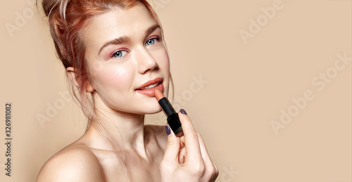 Portrait of gorgeous smiling woman holding rosy lipstick in hand. Model posing in studio and looking in camera gracefully. Beauty and cosmetics concept. Isolated on beige photo