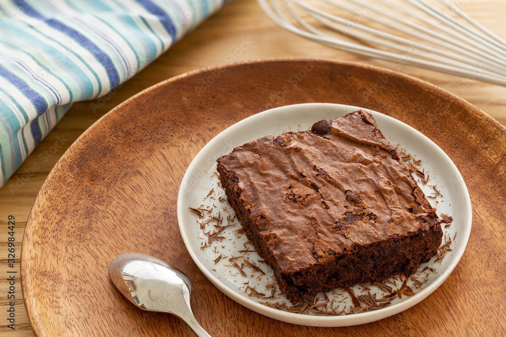 Brownies cake in a light gray plate with grated chocolate and silver spoon in a wooden plate by a white whisk over an oak table background. Brownies cake in a light gray plate with grated chocolate and silver spoon in a wooden plate by a white whisk over an oak table background.