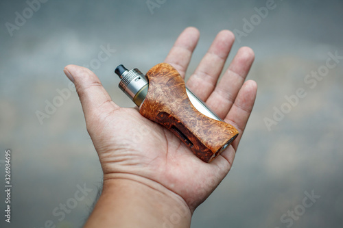 high end titanium genesis atomizer with stabilized natural black ash wood regulated box mods in hand on grey texture background, vaping device, selective focus photo