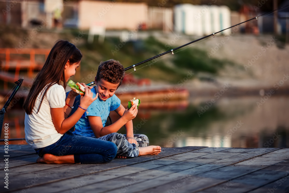 Two happy little friends, boy and girl talking, eating sandwiches and fishing on a lake Two happy little friends, boy and girl talking, eating sandwiches and fishing on a lake