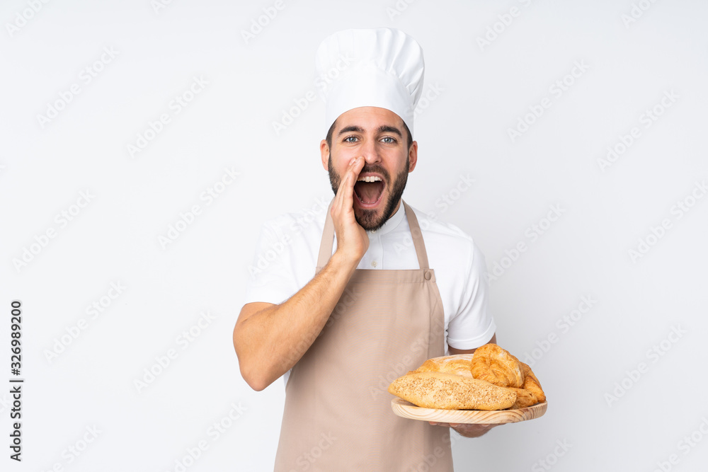 Male baker holding a table with several breads isolated on white background shouting with mouth wide open Male baker holding a table with several breads isolated on white background shouting with mouth wide open