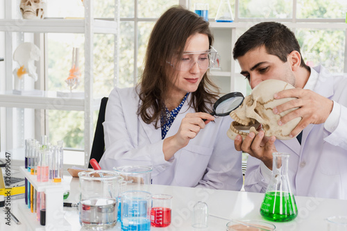 A male scientist is carrying a human skull while a female scientist is holding a magnifying glass on a skull in a science lab surrounded by a solution used in scientific experiments.