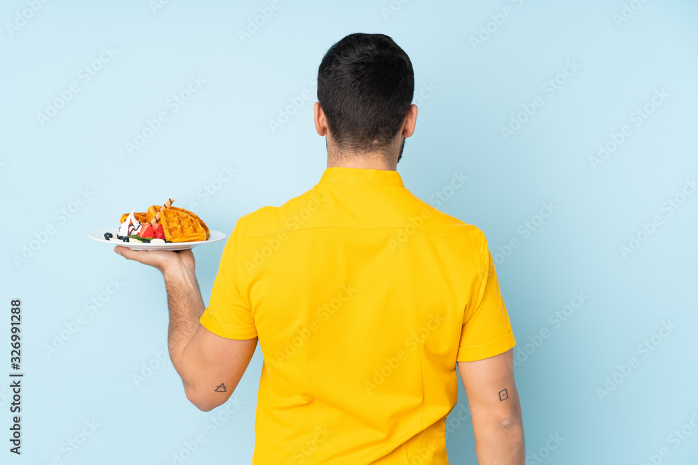 Caucasian man holding waffles isolated on blue background in back position