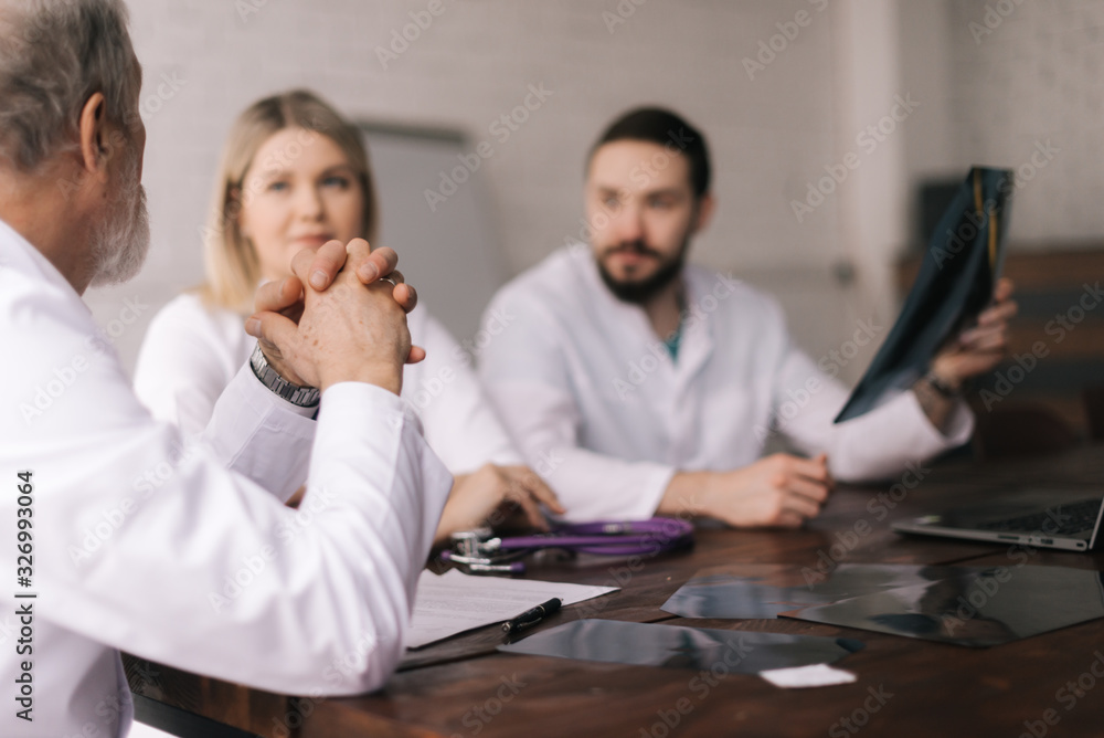 Team of three adult and young medical specialists discuss chest X-rays in the conference room of the hospital. Concept of team of doctors working together at the clinic. Concept of team medical work. Team of three adult and young medical specialists discuss chest X-rays in the conference room of the hospital. Concept of team of doctors working together at the clinic. Concept of team medical work.