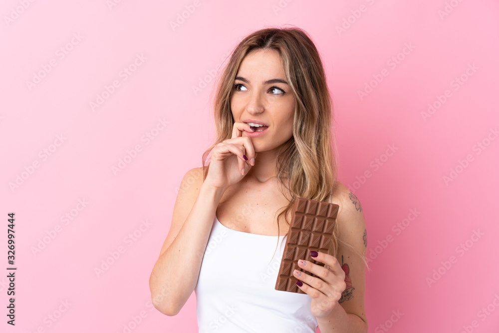 Young woman over isolated pink background taking a chocolate tablet and having doubts Young woman over isolated pink background taking a chocolate tablet and having doubts