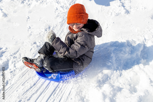 A teenager flies on a round sled from a hill. White snow scatters to the sides. Winter games for children. A Smiling boy with spectacles on eyes in orange hat riding down with extreme speed.Sunny day