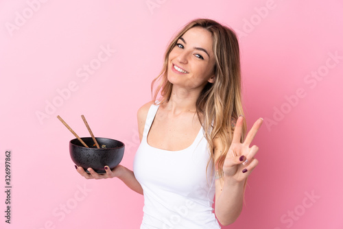 Young woman over isolated pink background smiling and showing victory sign while holding a bowl of noodles with chopsticks
