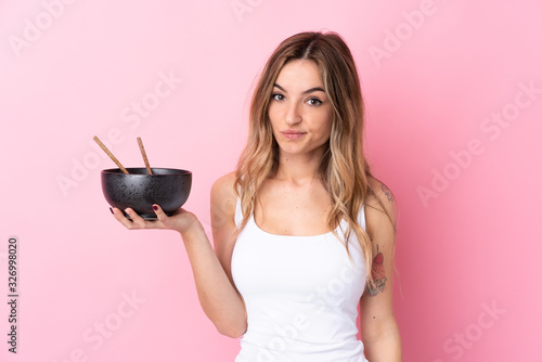 Young woman over isolated pink background with sad expression while holding a bowl of noodles with chopsticks