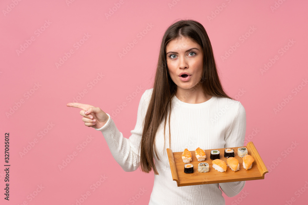 Young girl with sushi over isolated pink background surprised and pointing side Young girl with sushi over isolated pink background surprised and pointing side