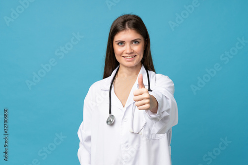 Young woman over isolated blue background with doctor gown and with thumb up