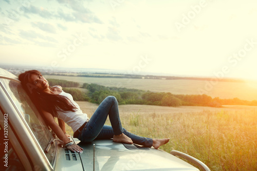Attractive yong woman is lying on the car's hood and looking at sunset. Rural evening background. © diter