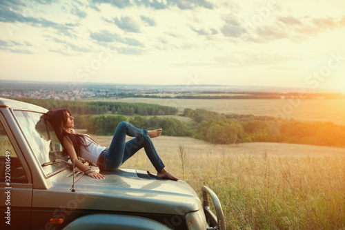 Attractive yong woman is lying on the car's hood and looking at sunset. Rural evening background.