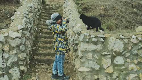 A boy in a jacket and hat takes pictures of a black cat sitting on a stone parapet photo