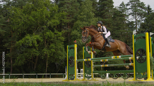 Young woman rider jumping over oxer fence on strong brown horse