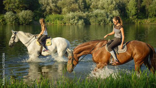 CLOSE UP: Two young girls riding horses along grassy river bank