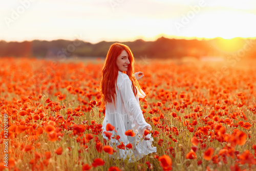 Happy redhead smiling woman in white dress on field of poppies at summer sunset