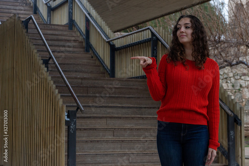 happy positive caucasian young woman model smiling pointing aside with index finger and looking aside, having great idea, in the park, orange sweater and jeans, long curly hair. Place for your text in