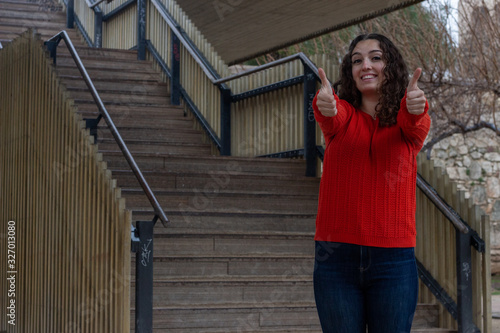 Portrait of happy confidence caucasian young woman model showing thumbs up gesture, in the park, orange sweater and jeans, long curly hair. Place for your text in copy space.