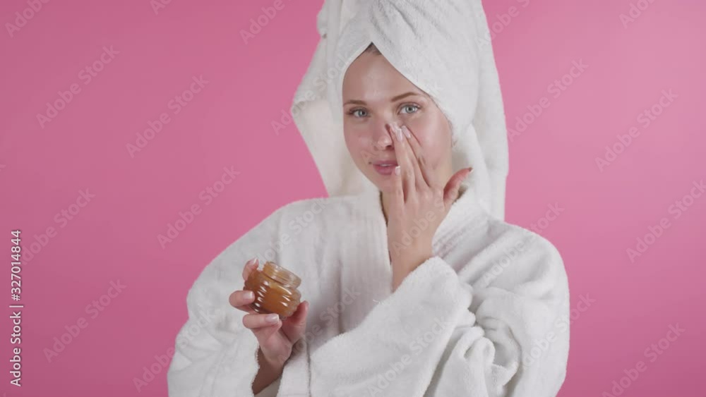 Portrait shot of beautiful young woman in shower robe and towel standing against pink background and applying moisturizing lotion to her face while smiling for camera Portrait shot of beautiful young woman in shower robe and towel standing against pink background and applying moisturizing lotion to her face while smiling for camera
