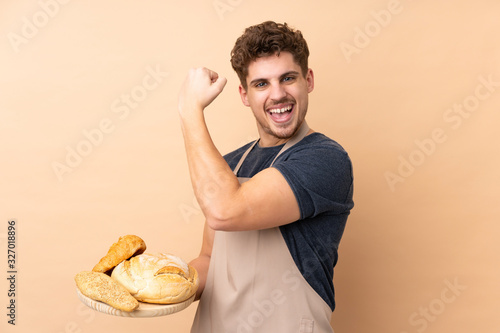Male baker holding a table with several breads isolated on beige background making strong gesture