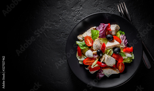 Fresh salad with chicken breast, cheese, black olives,red pepper, lettuce, fresh sald leaves and tomato on a black plate on Dark grey black slate background