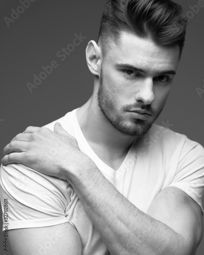 Monochrome portrait of a young man with a beard on a gray background. A muscular man in a t-shirt. Studio portrait. Fitness, fashion and beauty
