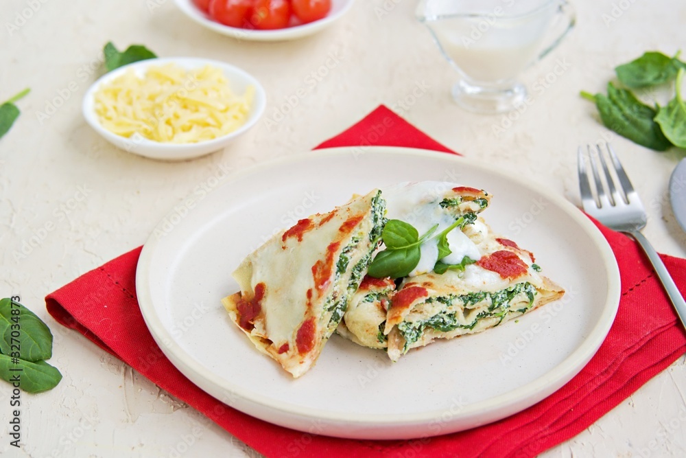 Crepes with spinach and ricotta cheese, baked with bechamel sauce and tomato sauce on a white plate on a light concrete background. Easter Italian food. Italian food. Crepes with spinach and ricotta cheese, baked with bechamel sauce and tomato sauce on a white plate on a light concrete background. Easter Italian food. Italian food.