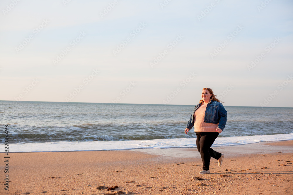Beautiful overweight woman running on the sandy beach. Plus size girl enjoy warmth sunset with romantic mood. Fat model dressed jeans jacket and pink knitted sweater Beautiful overweight woman running on the sandy beach. Plus size girl enjoy warmth sunset with romantic mood. Fat model dressed jeans jacket and pink knitted sweater