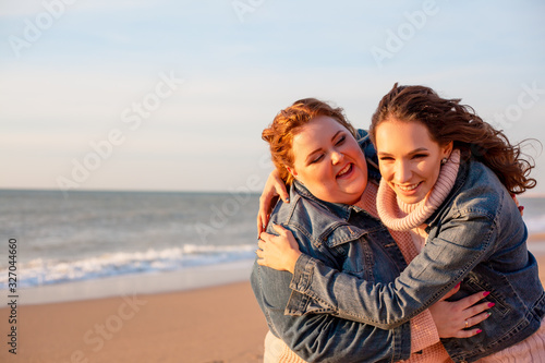 Back view of two freinds   plus size with  thin  girls walking on the spring beach. Fat woman with strong friend  laughting. Overweight woman dressed jeans jacket and pink sweater. 