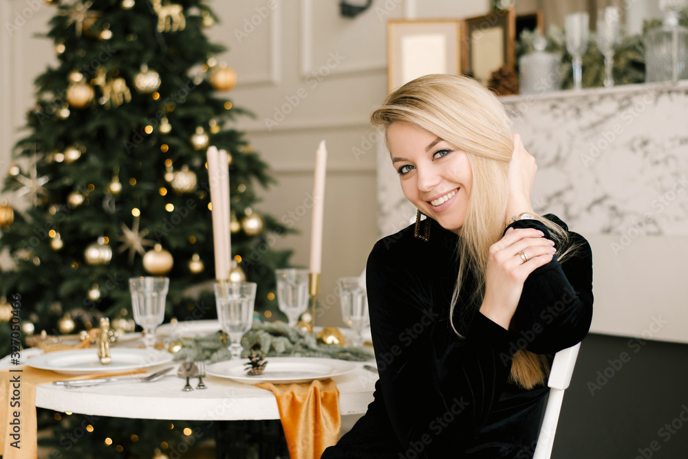 Christmas portrait of a smiling blonde girl in a black dress on the background of Christmas decor at a table in an elegant interior. A woman is preparing to celebrate Christmas and New year