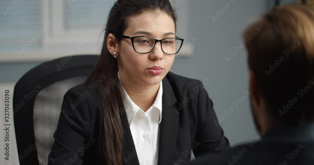 Brunette female employer in white shirt and black suit sitting at table in own office and asking question male employee. Beautiful woman in eyeglasses interviewing new candidate Brunette female employer in white shirt and black suit sitting at table in own office and asking question male employee. Beautiful woman in eyeglasses interviewing new candidate