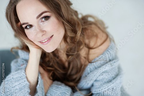 Close-up portrait of attractive young European smiling girl with light brown hair in light blue clothes. She poses sitting on a light blue chair. Studio shooting. Light background.