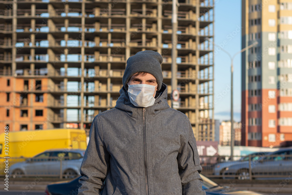 a guy is standing in the city near the road. Around unfinished apartments, traveling cars. Wearing a protective medical mask on his face. Danger of walking their viruses, coronavirus and diseases.