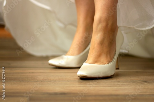 A portrait of the feet of a bride wearing white high heeled wedding shoes while pulling her dress up to show them. The woman is standing on a wooden floor with her high heels ready to get married.