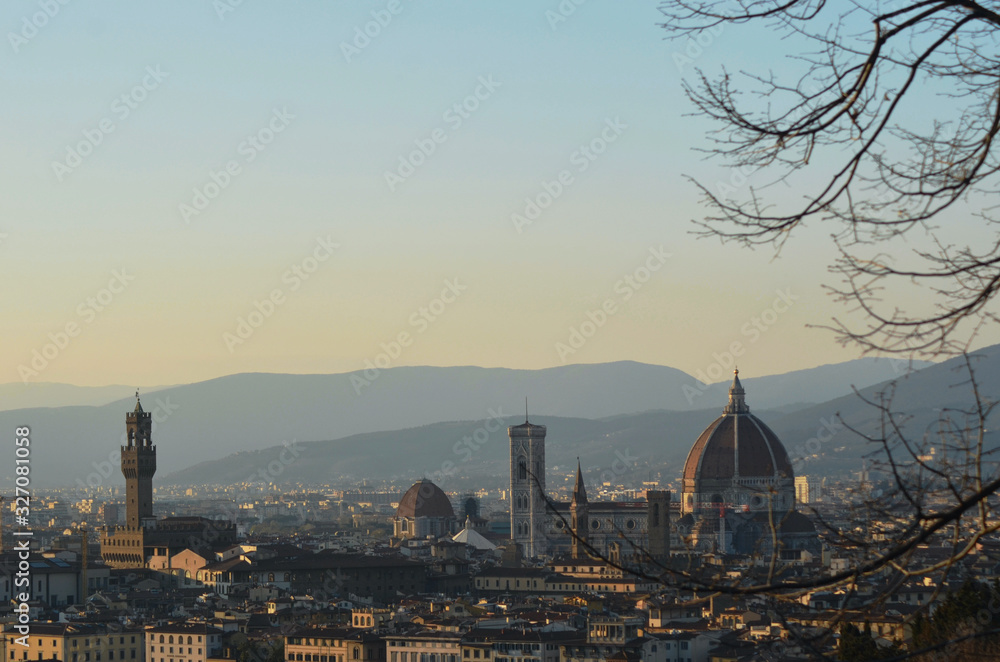 The bare branches of a tree frame the view over the city of Florence, Italy at dusk. The Duomo, Baptistery and Campanile dominate the skyline. Hills are in the background. The sky is clear.