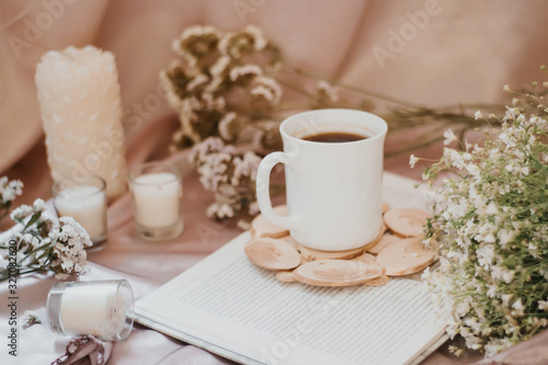 A cup of coffee on a stand surrounded by flowers and candles of different sizes and shapes