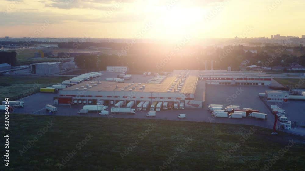 Aerial side view of the logistics park with warehouse, loading hub with many semi trucks with cargo trailers standing at the ramps for load/unload goods at sunset Aerial side view of the logistics park with warehouse, loading hub with many semi trucks with cargo trailers standing at the ramps for load/unload goods at sunset