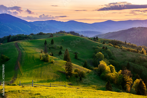 mountainous countryside in springtime at dusk. road  wooden fence and trees on the rolling hills. ridge in the distance. clouds on the sky. beautiful rural landscape of carpathians