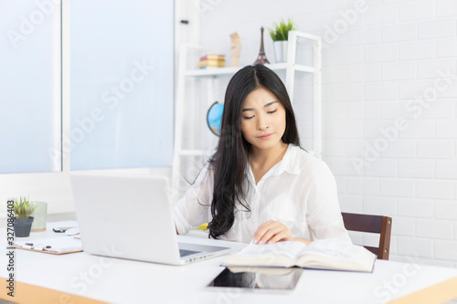 Education study abroad, Overseas Asian student girl sitting at table looking book while do homework by laptop making video call abroad to internet friend connection, happy mood smile broadly library