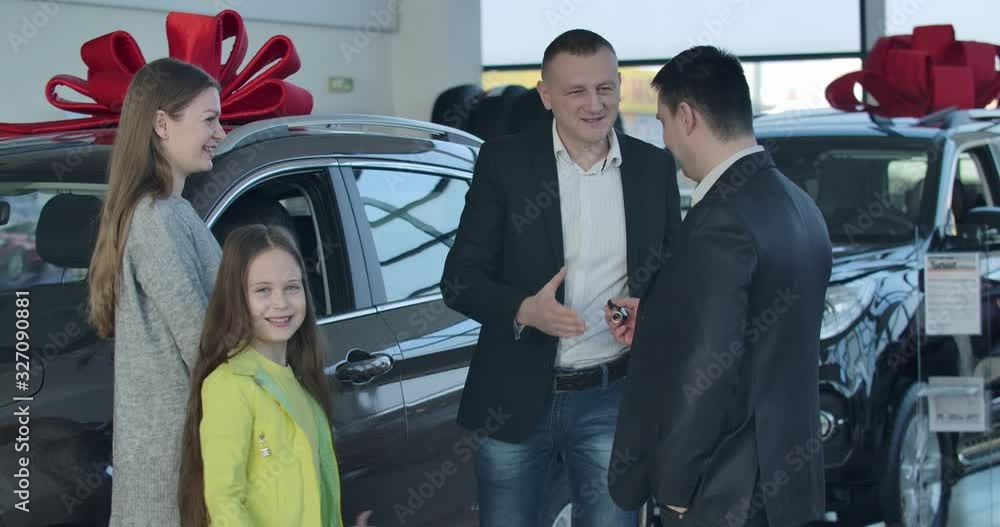 Positive Caucasian dealer giving car keys to man, shaking clients' hands. Cheerful girl showing victory gesture to camera. Happy young family buying automobile in dealership. Cinema 4k ProRes HQ. Positive Caucasian dealer giving car keys to man, shaking clients' hands. Cheerful girl showing victory gesture to camera. Happy young family buying automobile in dealership. Cinema 4k ProRes HQ.